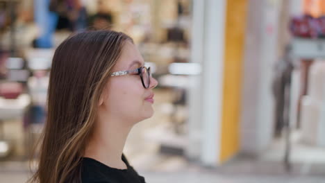 confident woman in glasses walking through a modern shopping mall with blurred background lights and store interior, stylish young shopper enjoying a relaxed shopping