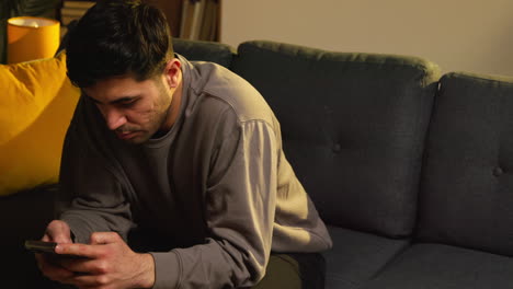 young man spending evening at home sitting on sofa with mobile phone scrolling through internet or social media 11