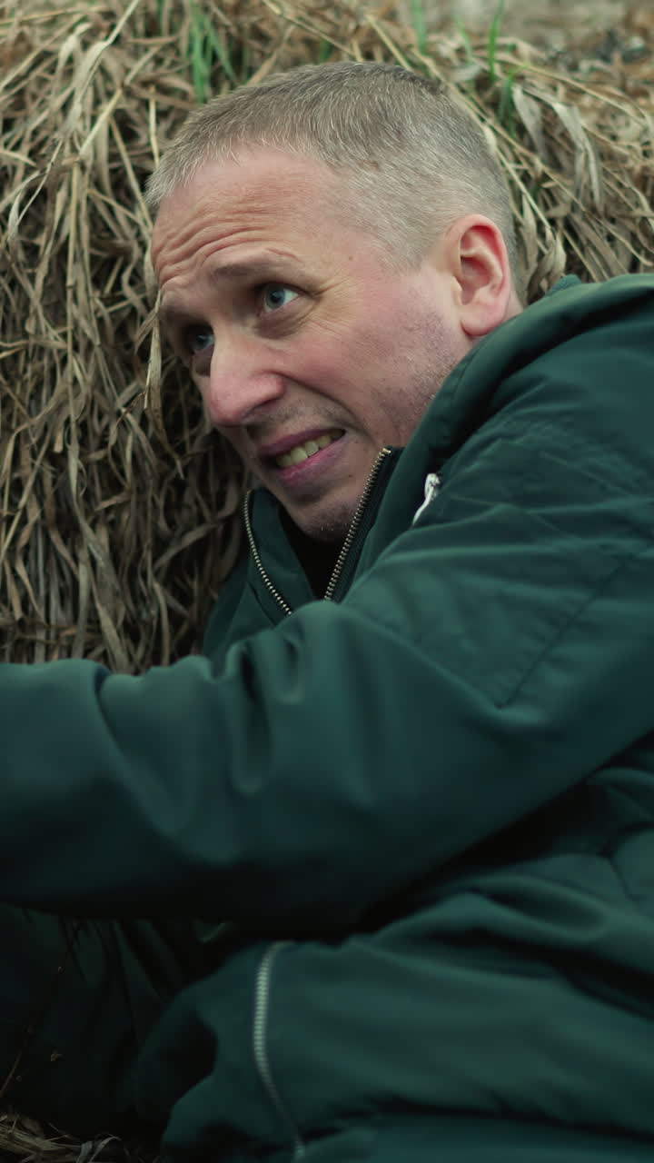 una vista cercana de un hombre con chaqueta verde y vaqueros azules sosteniendo una pistola, mirando y tenso mientras se acuesta contra la hierba seca