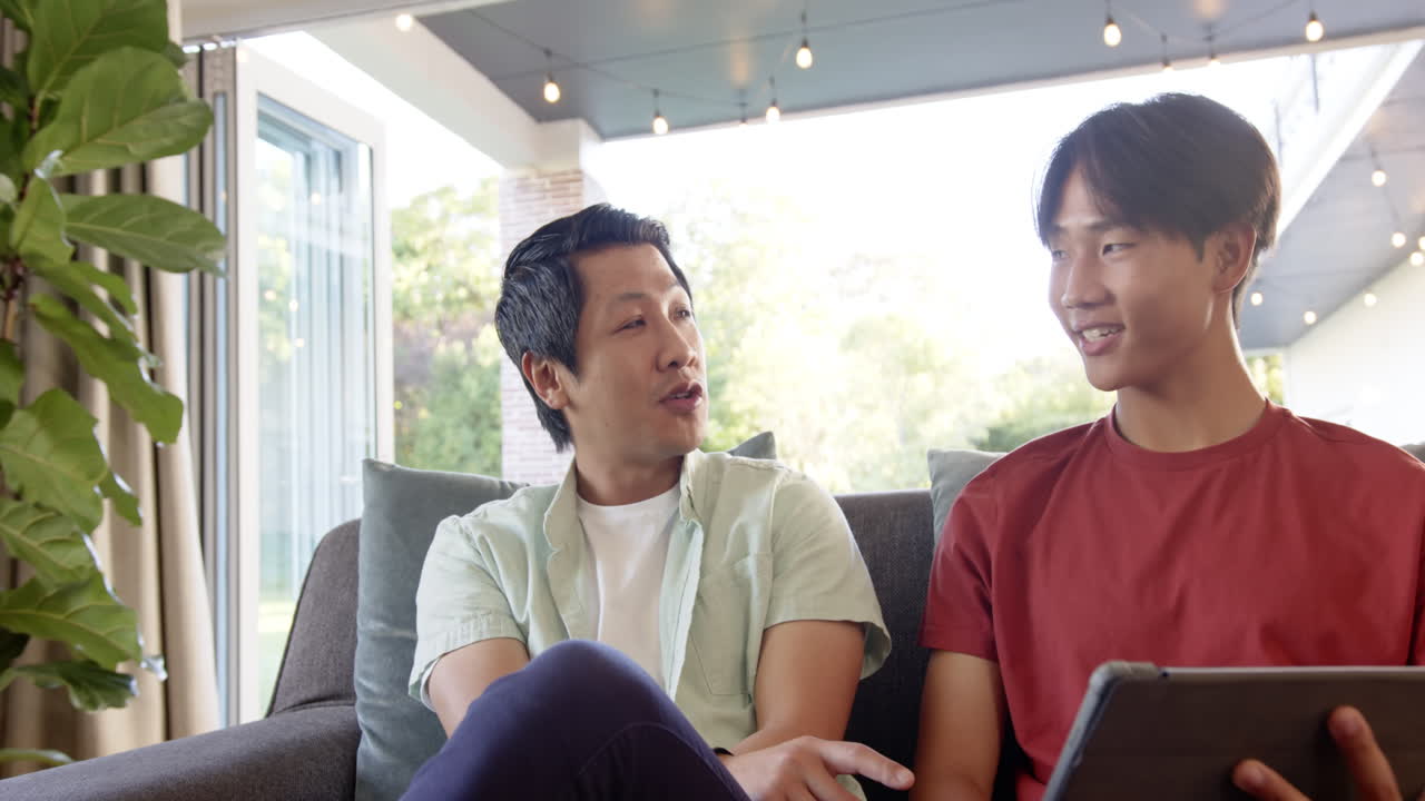 Asian father and teenage son sitting on couch, smiling and interacting with a tablet
