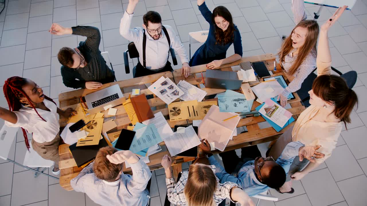 Set ovenfra: Glad gruppe forretningsfolk arbejder sammen, samler hænderne, klapper og fejrer succes på et moderne kontor.