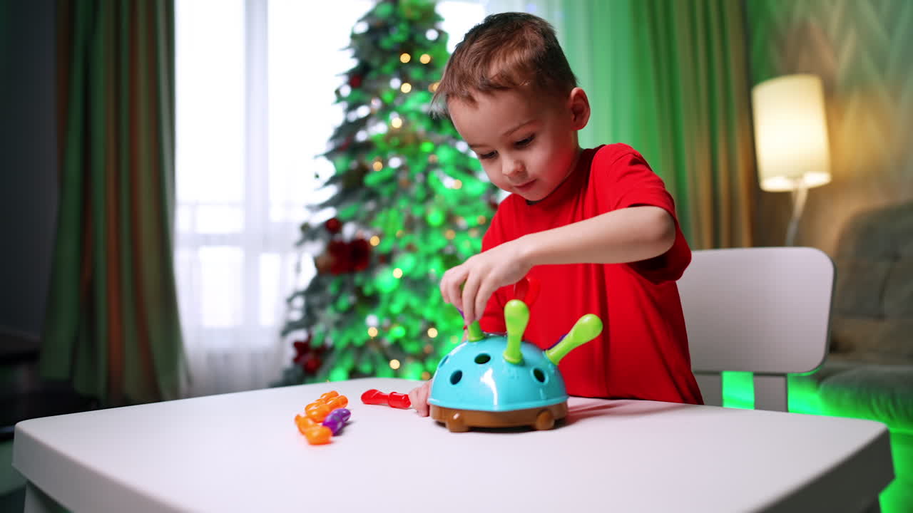 Beautiful Caucasian toddler in red t-shirt stands at desk playing with toy. Baby playing at home. Christmas tree at backdrop.
