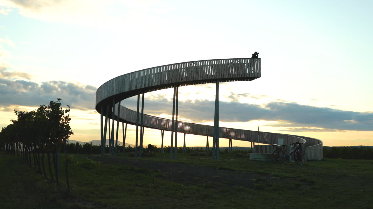 mirador edificio giratorio de madera con figura gris de hombre con binoculares mirando el horizonte en el fondo nubes en movimiento y sol poniente durante la noche de otoño zona del sur de moravia