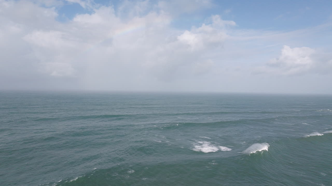 Aerial drone shot of big Atlantic Ocean waves coming into shore on a day with giant waves in Nazaré, Portugal, Europe. Drone flying towards rainbow and waves, sunny with clouds