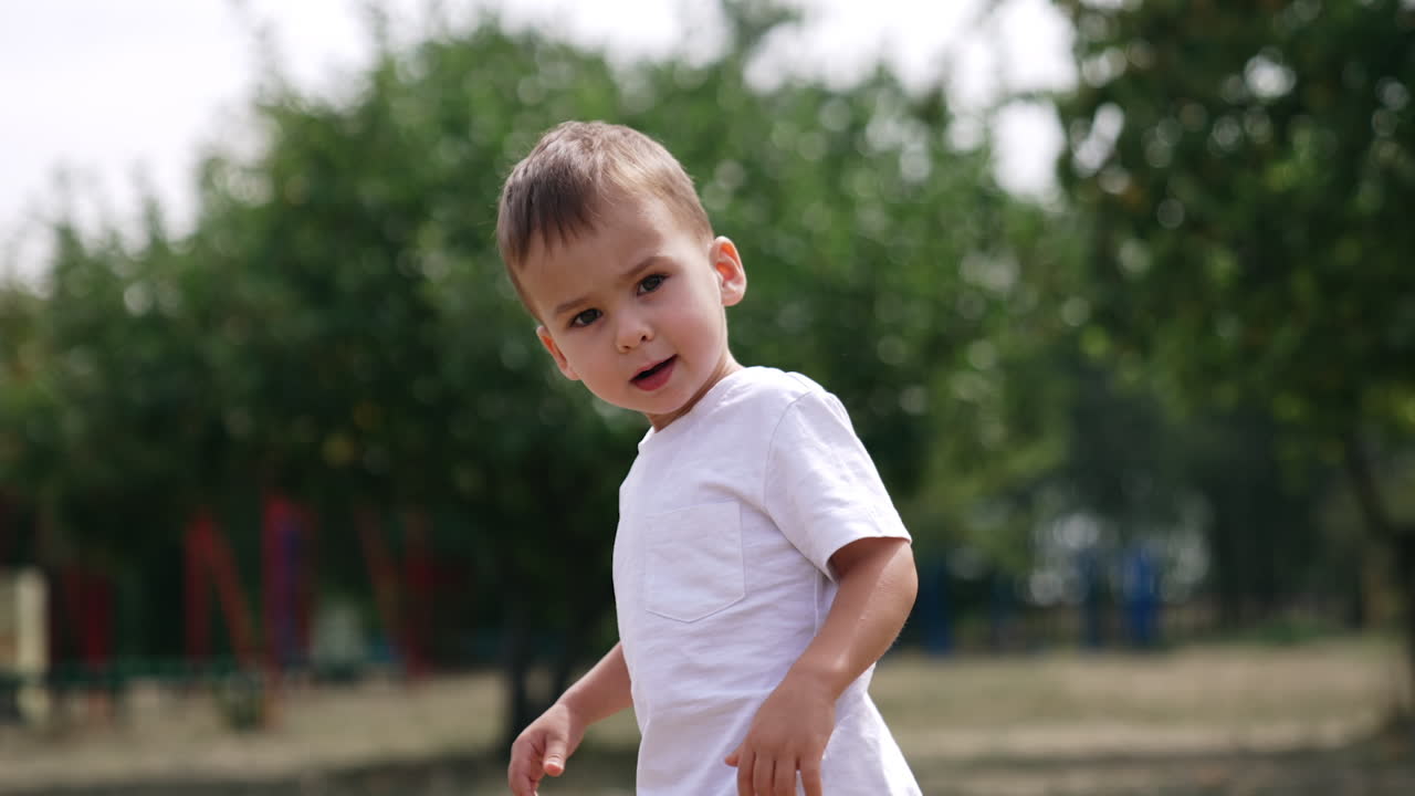 Cute Caucasian baby boy in white t-shirt stands looking at camera turning his head to the side. Lovely child wants to walk away but turns and smiles.