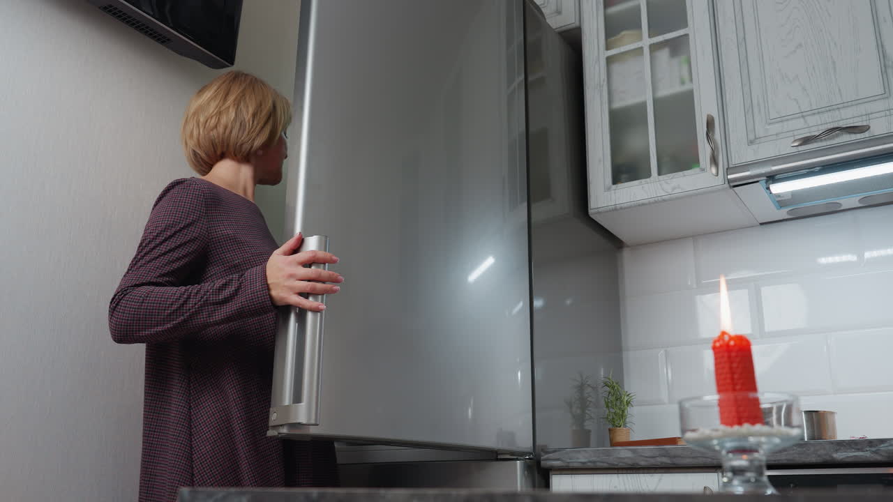 mujer abre la nevera para recuperar una caja de huevos mientras está de pie cerca de un enchufe de pared y la televisión, luego cierra la nevera, se aleja, y coloca los huevos en la encimera en una cocina moderna