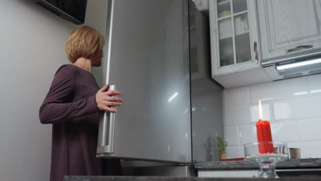 mujer abre la nevera para recuperar una caja de huevos mientras está de pie cerca de un enchufe de pared y la televisión, luego cierra la nevera, se aleja, y coloca los huevos en la encimera en una cocina moderna
