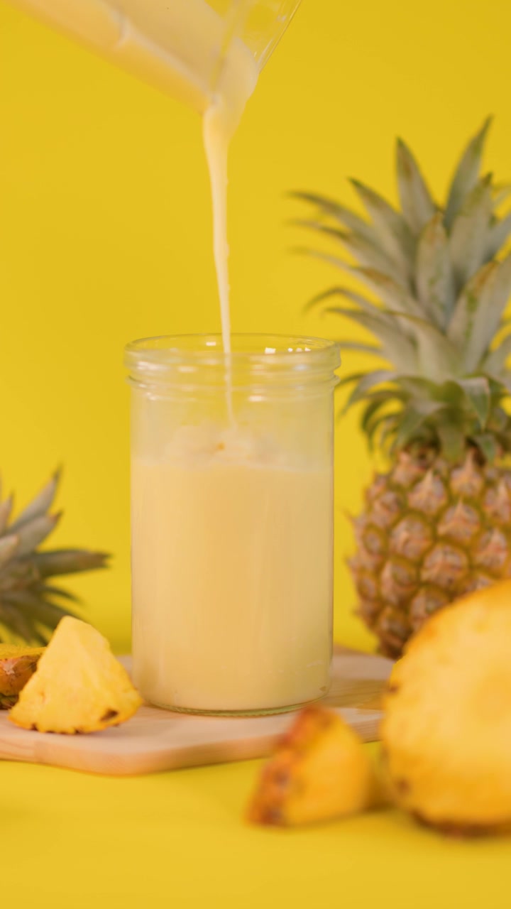 Smoothie all'ananas versato in un bicchiere, circondato da frutta fresca, su sfondo giallo brillante e con illuminazione soffusa.