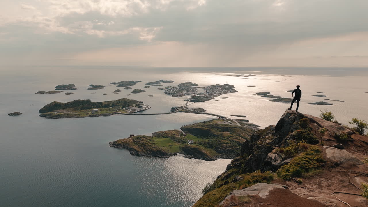 Aerial view over Henningsvær from Festvågtind with a man standing towards the small islands in summer. 4k high quality cinematic drone view