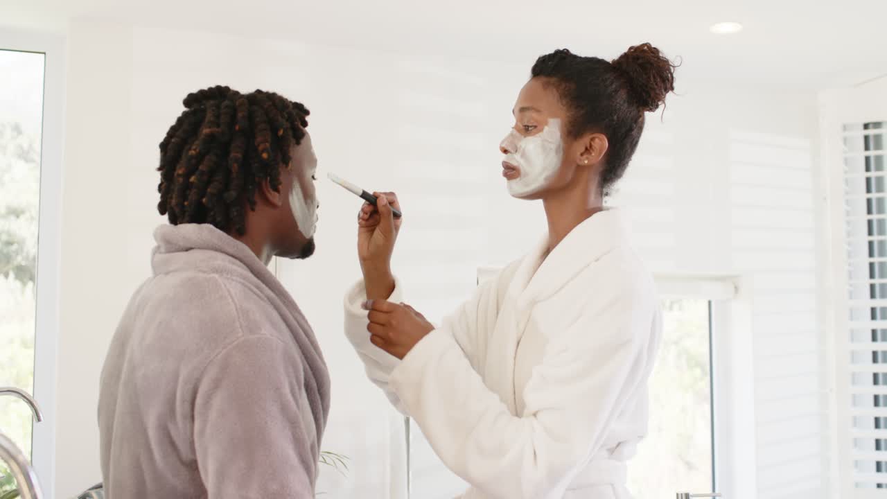 African American couple at sink, woman dipping brush in jar, applying mask on cheek for skincare