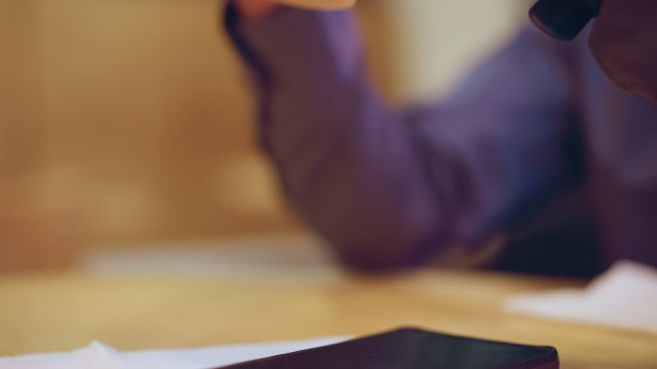 Close-up of hand in smart shirt holding takeaway coffee cup above wooden table with smartphone resting on napkin, soft background blur conveying moment of pause, tech presence, and casual indoor ambiance