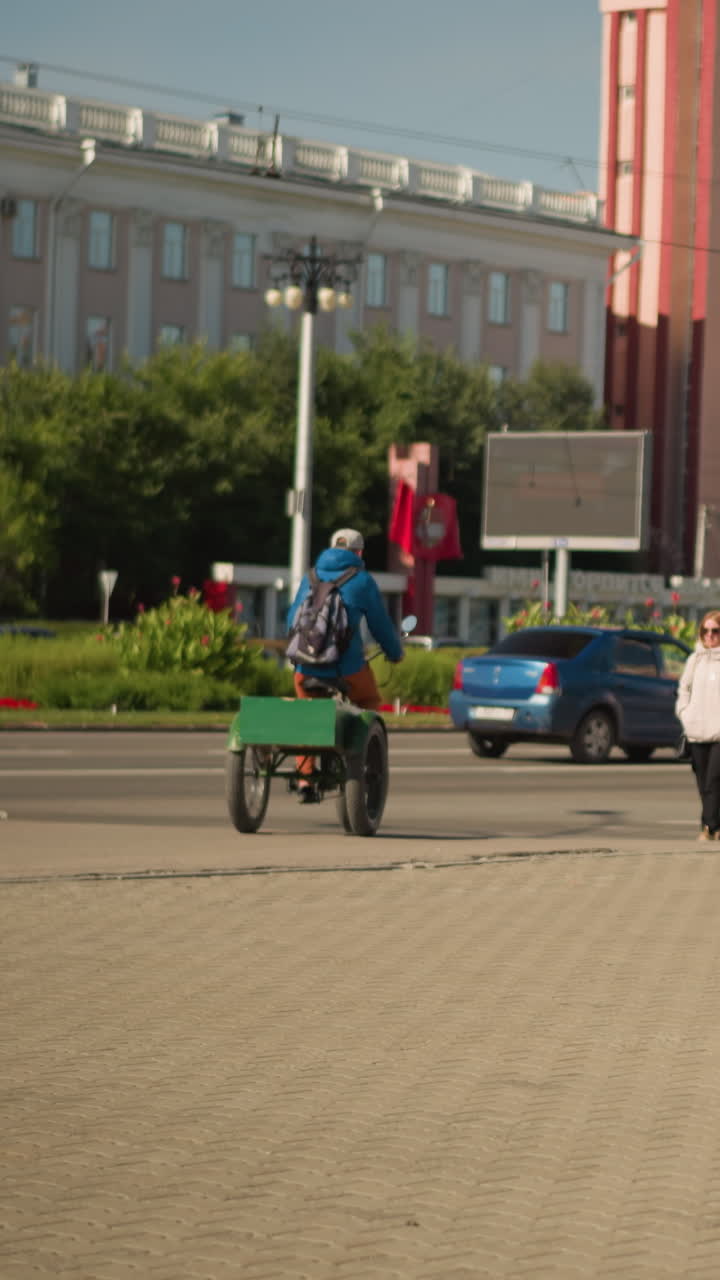 파란색 재킷과 백팩을 착용한 사람이 화물용 세발자전거를 타고 광장을 가로지르는 모습이에요. 배경에는 자동차와 광고판이 있는 녹색 화물 상자가 보이고, 여름 낮 시간의 도시 광장에서 택배 기사가 교통 체증을 헤쳐나가며 라스트마일 딜리버리를 하고 있어요.