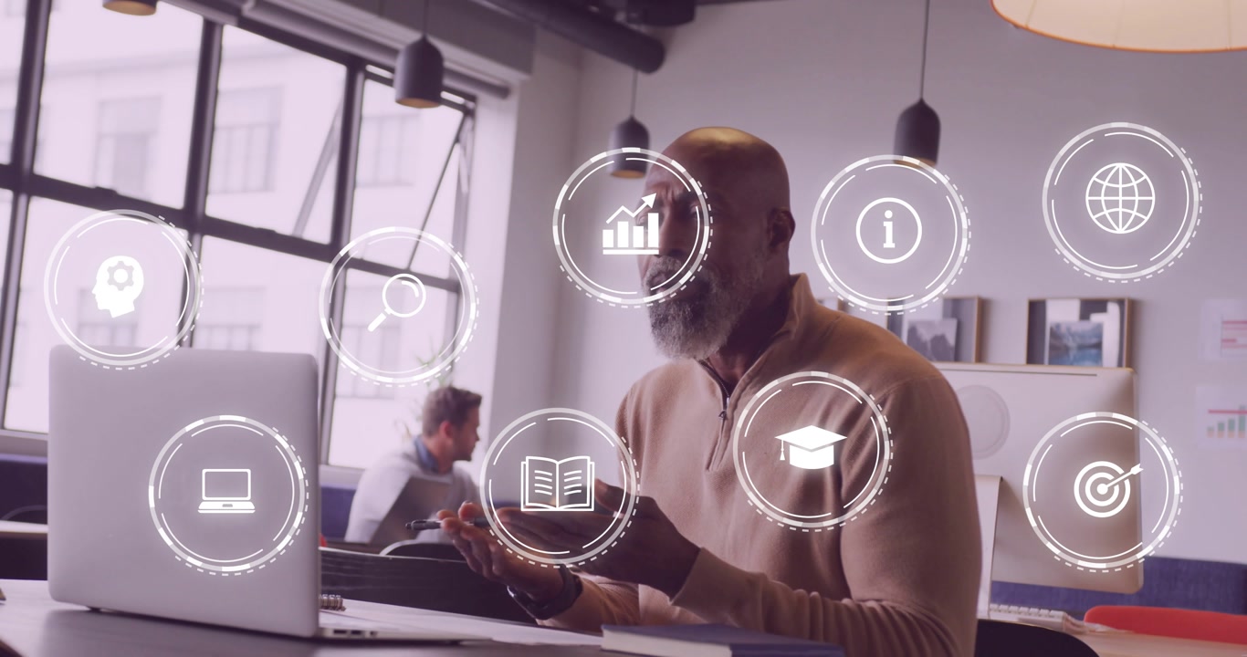 Consultant gesturing at laptop in open office, causing floating technology icons and taking notes