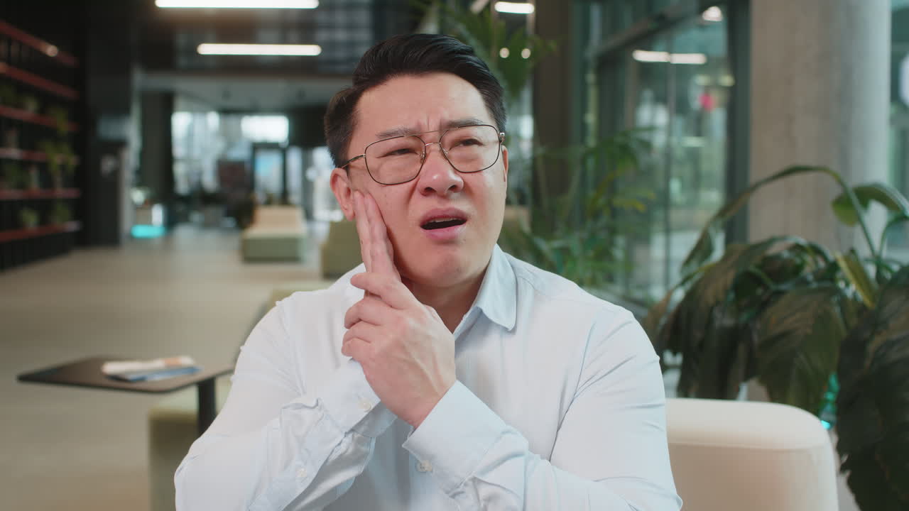 Chinese businessman in office lobby working on laptop and feeling toothache in modern office lobby