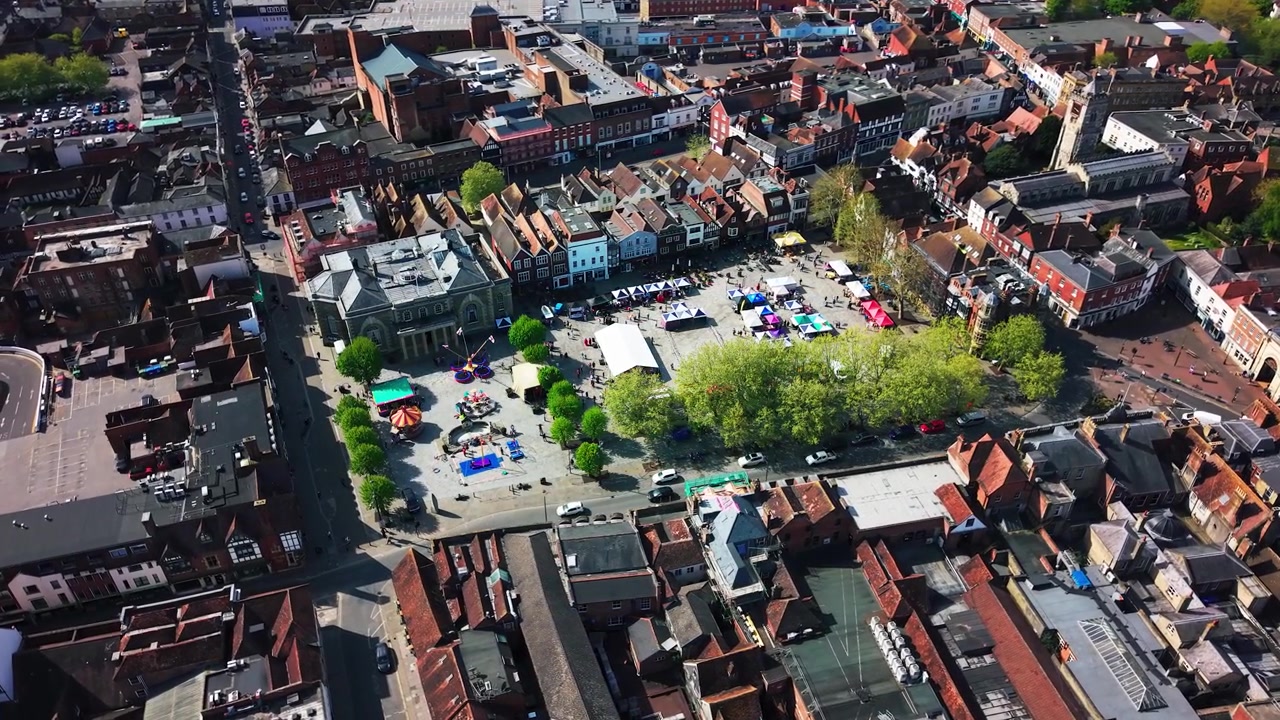 Aerial drone footage of a St George’s Day event taking place in Salisbury Market Square, showing stalls, crowds, historic buildings, streets, rooftops, and the city centre from above.