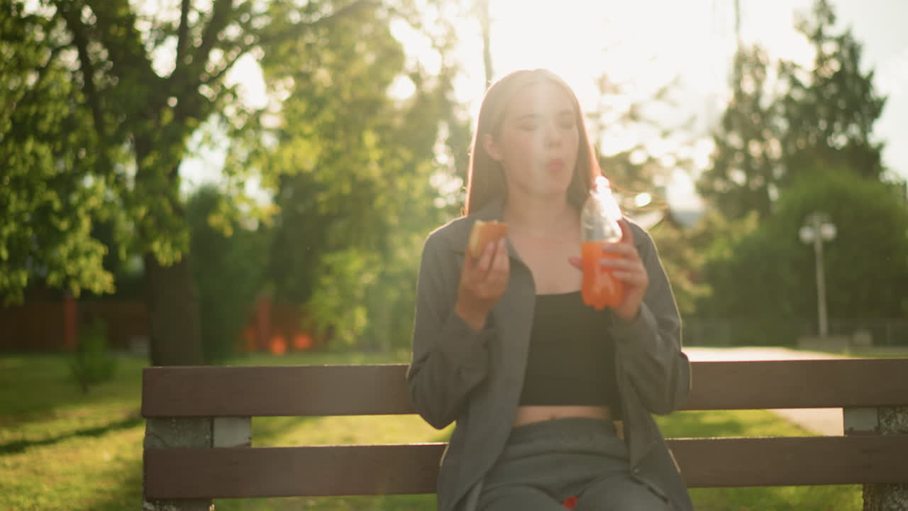 회색 옷을 입은 여자가 야외에 공원 벤치에 앉아 간식을 먹고 조용한 공원 환경에서 주스 음료 한 병을 들고, 그녀는 울창한 초록색과 나무로 둘러싸여 있습니다.