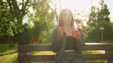 lady dressed in grey sitting on park bench outdoors, eating a snack and holding a bottle of juice drinks from it, in a tranquil park setting, she is surrounded by lush greenery and trees