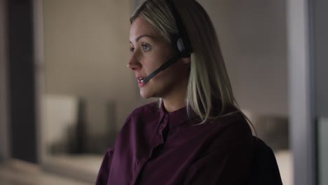 Video-portrait-of-smiling-caucasian-businesswoman-talking-on-headset,-working-at-night-in-office