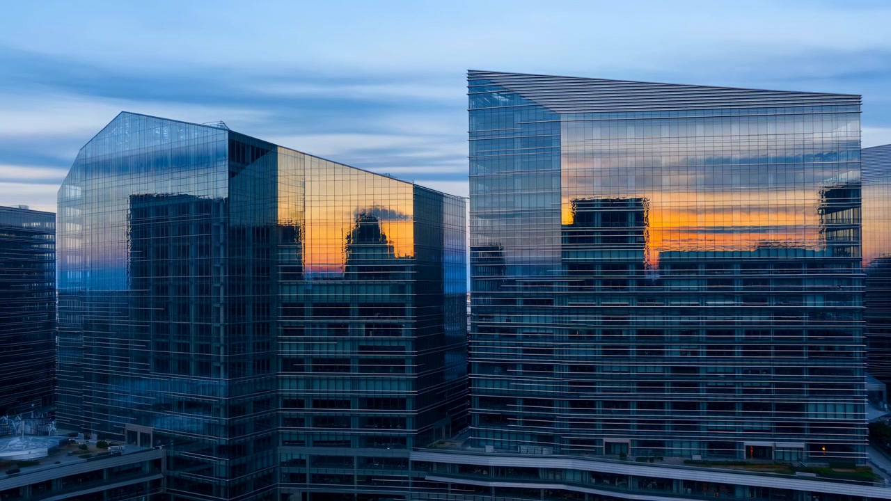 Glass office towers rise in a modern business district, reflecting warm golden sunset light across a calm urban skyline with contemporary architecture and mirrored facades