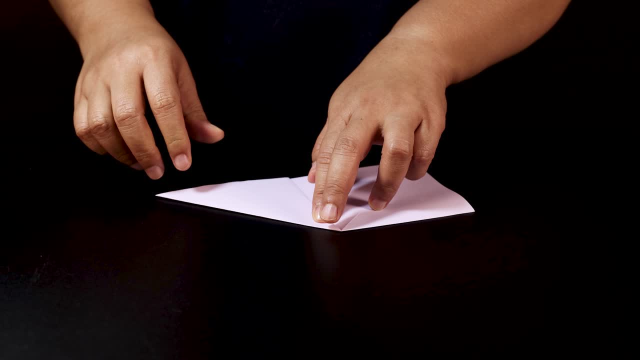 Adult hands methodically fold white paper on black table under soft, even lighting, overhead view