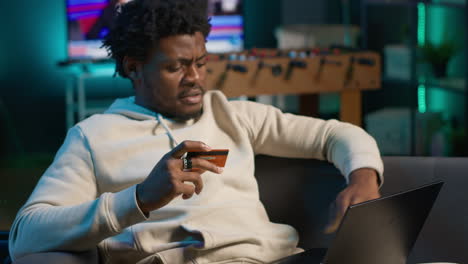 smiling african american man entering card digits to confirm a money transfer, setting up the payment for online shopping on his pc. happy person feeling good after placing orders. camera a.
