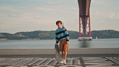 sportive guy posing skateboard at embankment. hipster standing river view place