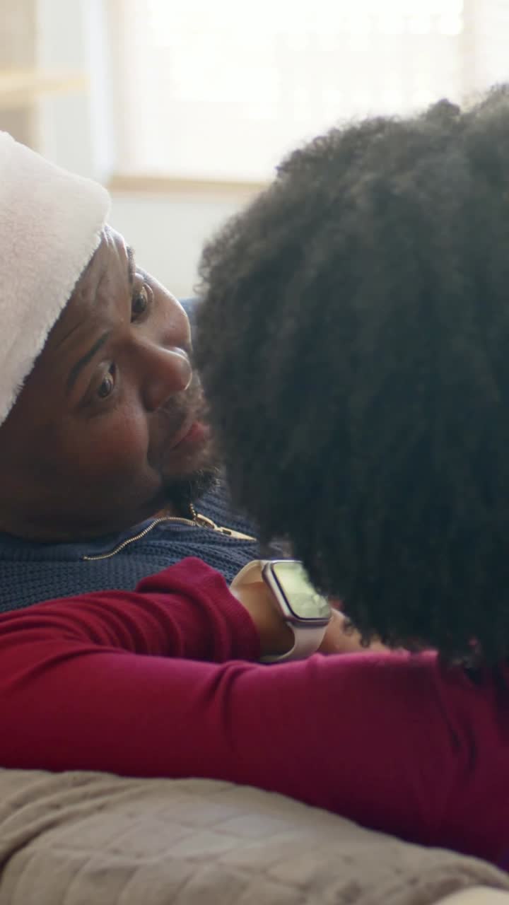 Video verticale: Un partner che si appoggia e mostra uno smartwatch a un uomo sul divano, suscitando un sorriso con un cappello da Babbo Natale.