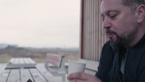 adult male drinking coffee under pergola in exterior while looking at smartphone with smile, slow motion