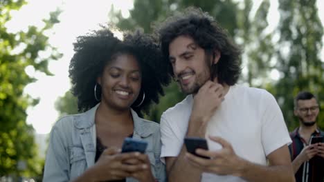 una pareja multirracial feliz usando teléfonos inteligentes en el parque.