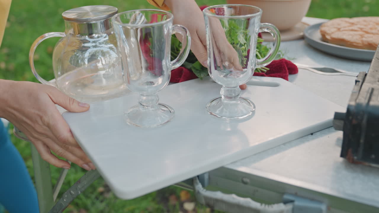 Woman setting camping table with glassware and picnic food in birch forest yellow jacket host carries tray with glasses, pitcher and salad to portable table, bread and plates arranged, calm daylight