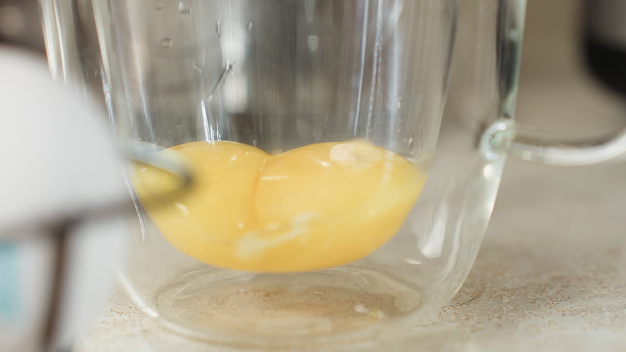 Close up of egg yolks inside glass cup with another yolk gently dropping in, showing slow careful movement of ingredient separation during kitchen prep for baking