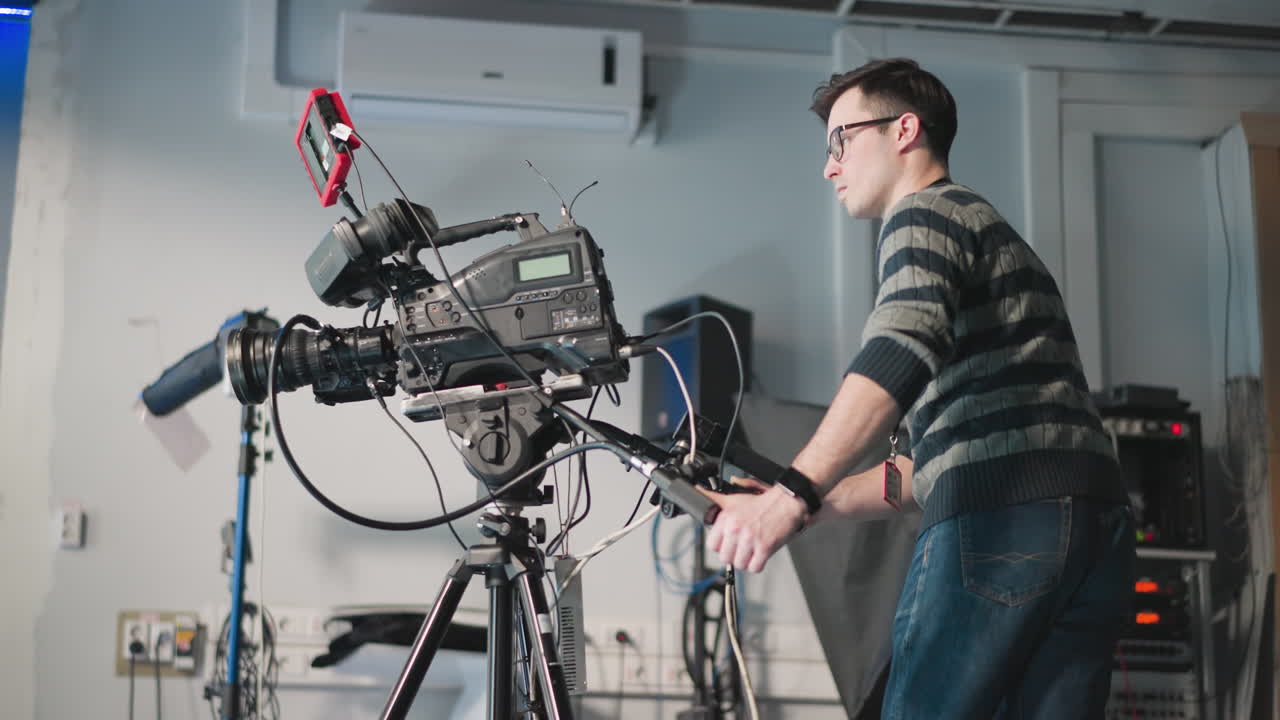 Man adjusting professional video camera on tripod in studio. Technician focused on camera setup with screen and equipment. Filming session ongoing in production environment