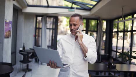 and adult man in a shirt walks through a restaurant with a laptop and speaks on the phone