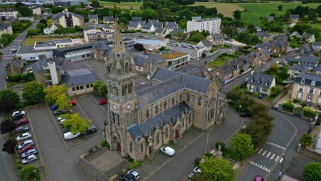 the drone orbits around saint-sauveur church in plancoët, showing parked cars, a passing bird, surrounding houses and green fields. brittany in france
