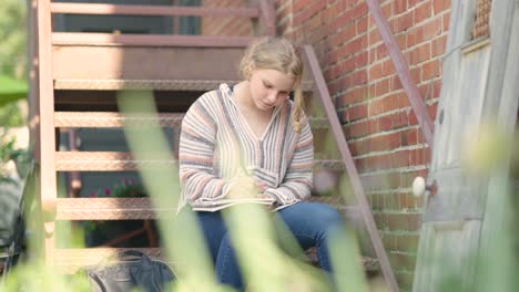 girl student writes on notepad while sitting on metal stairs outside