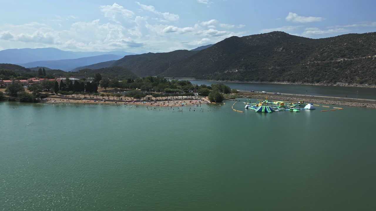Turquoise water of lac vinca in france, with tourists enjoying a sunny summer day. A large inflatable water park structure provides entertainment