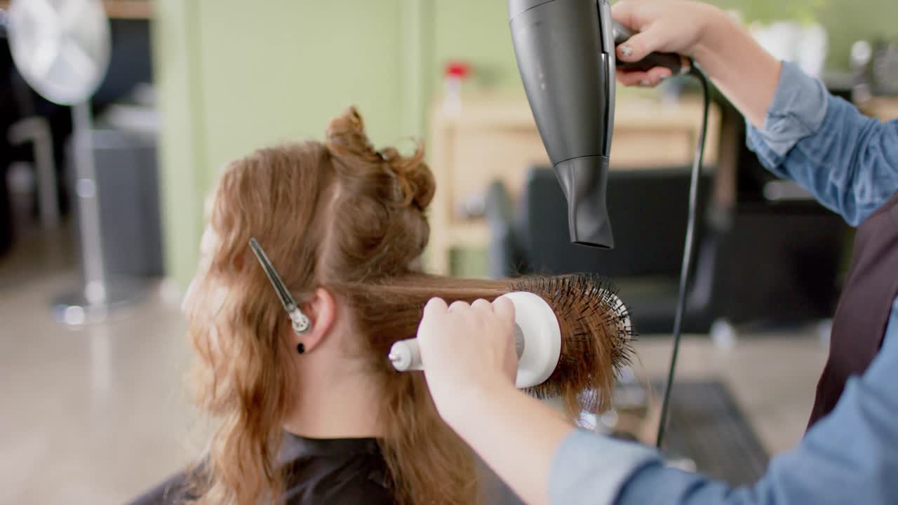 Caucasian female hairdresser styling male client's long hair with hairdryer and brush, slow motion