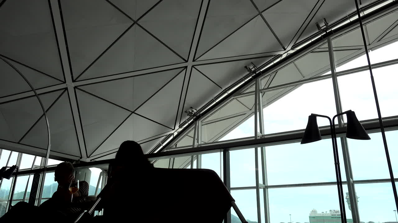 silhouette of a man and a woman sitting in a modern waiting lounge at the airport looking at their cell phones and drinking a glass of wine
