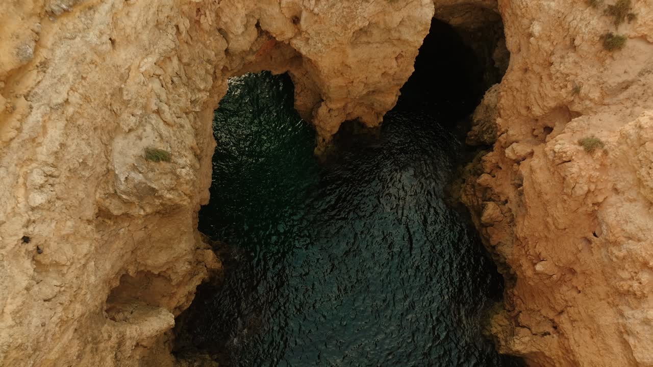Aerial shot rising above textured rock formations revealing a natural arch over deep turquoise water in Algarve