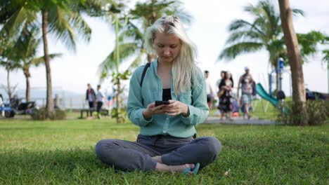 beautiful slim woman with long blonde hair in green shirt sits on the ground and using smartphone over background the park. girl on the square touching screen and smile