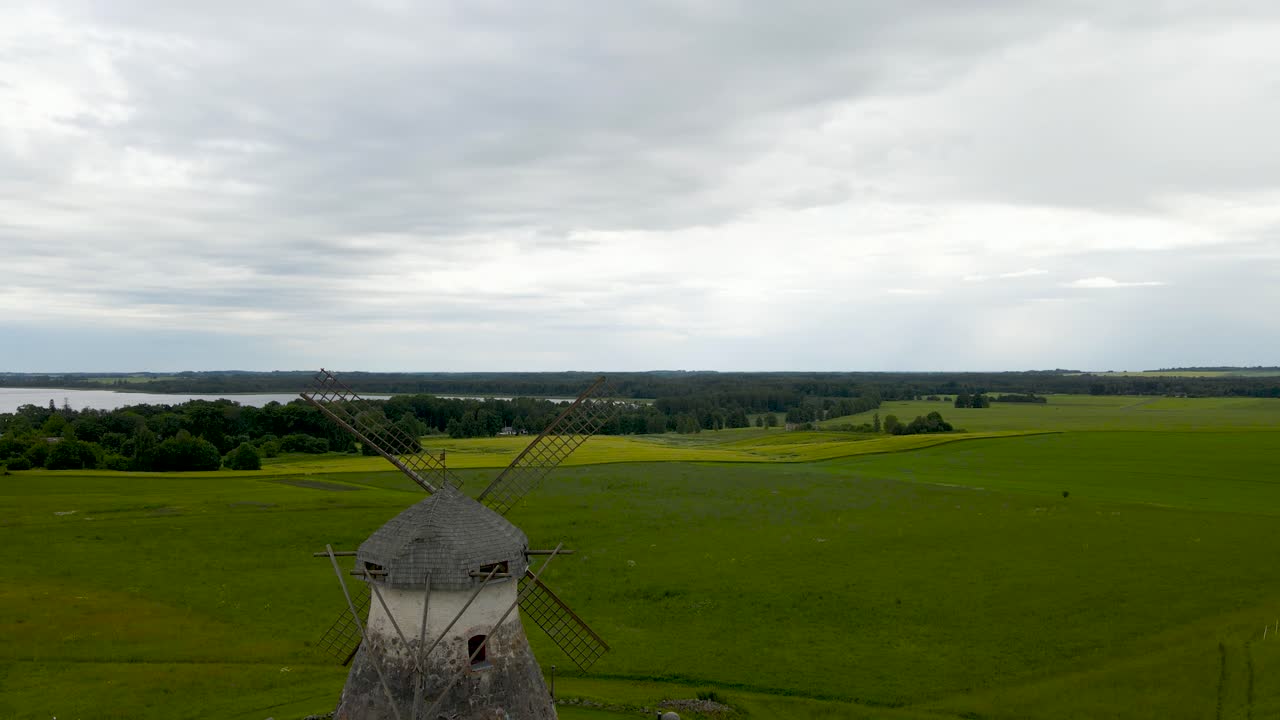 Drone glides forward above the rural landscape of Kuremaa, Jõgeva. Forward view of an old style stone windmill with classic wooden blades, revealing verdant farmland with tree lines and large lake