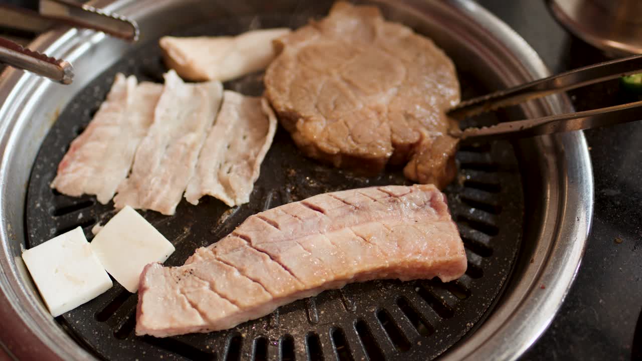 Hand uses tongs to flip pork slice on tabletop Korean barbecue grill under warm lighting