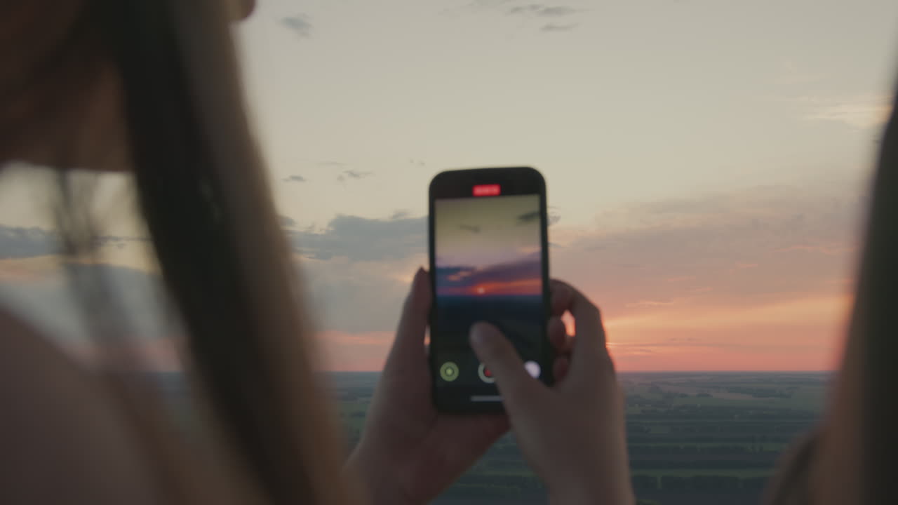 Close back view of woman holding smartphone to capture glowing sunset across wide open landscape, soft colors filling sky as warm light reflects on phone screen