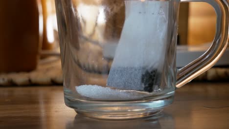 Taking-electric-ketle-and-pouring-water-inside-a-glass-cup-with-sugar-and-tea-bag-for-tea-preparation