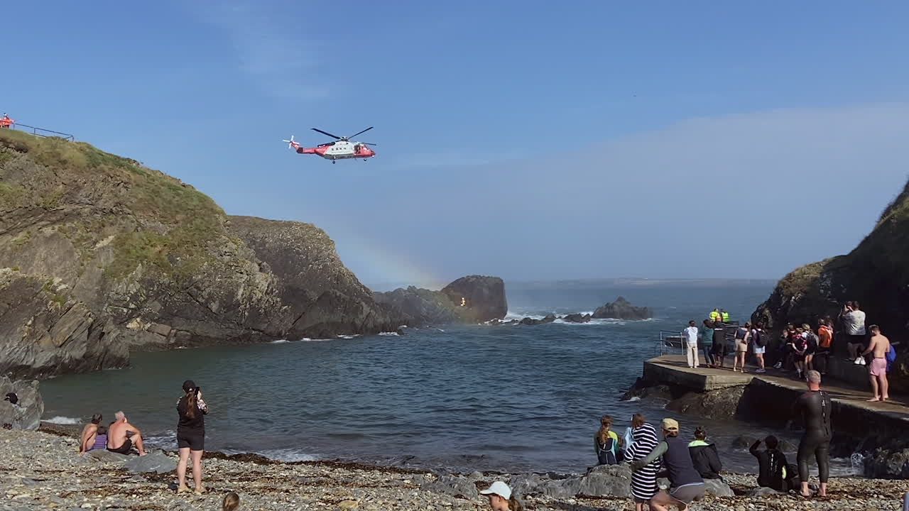 Coastguard helicopter lowers rescuer thru rainbow to rocky shoreline