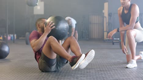 Entrenadora-Diversa-Motivando-A-Un-Hombre-Decidido-A-Hacer-Ejercicio-Con-Balón-Medicinal-En-El-Gimnasio,-En-Cámara-Lenta