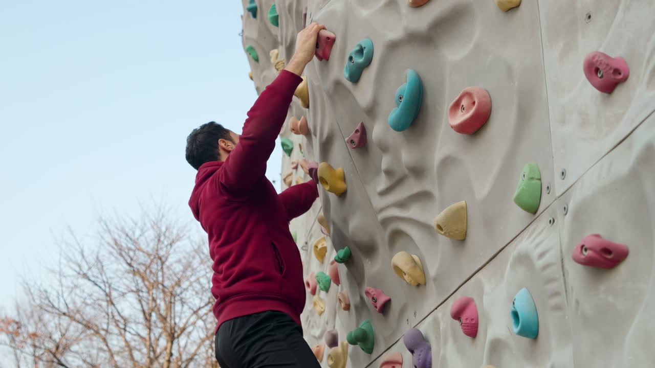 un hombre barbudo de treinta años sube a una pared de escalada en un campo deportivo al aire libre para fortalecer el agarre de la mano