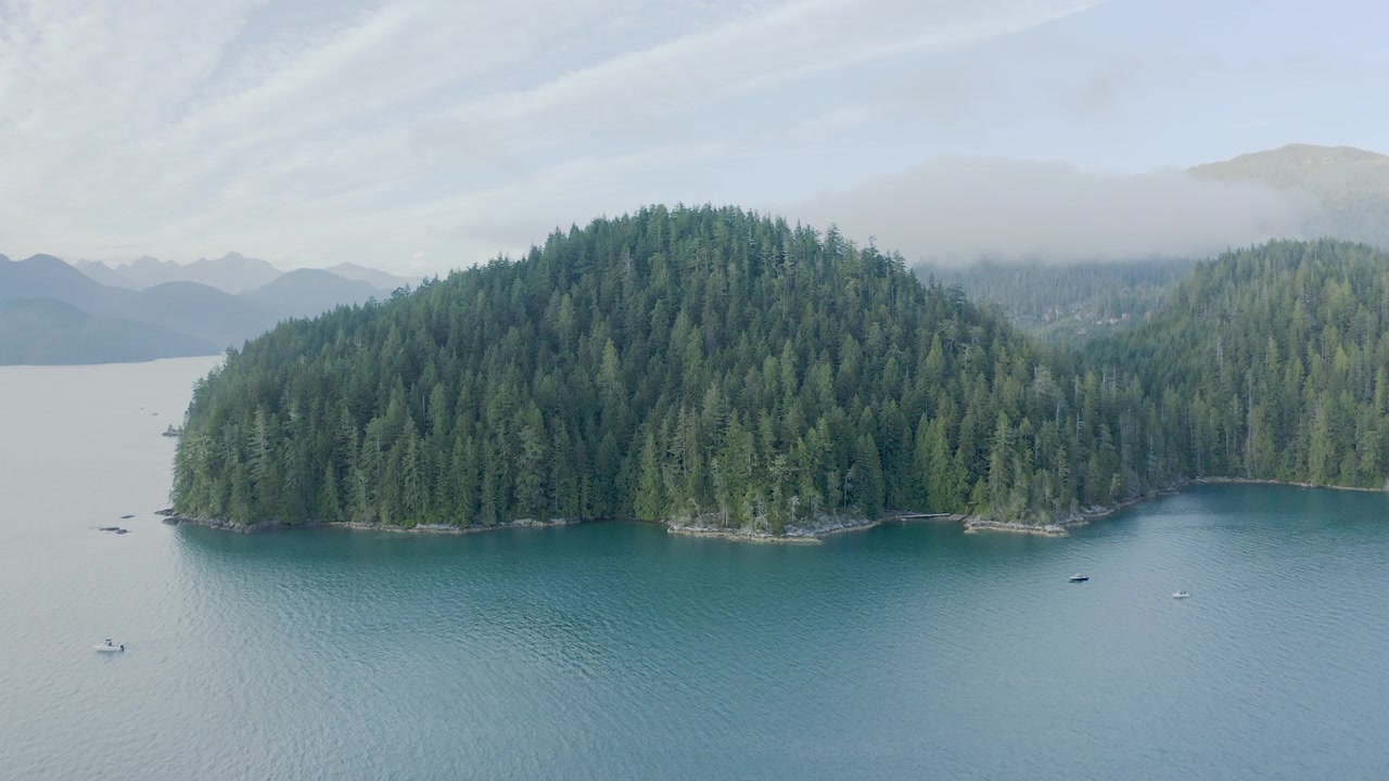 Un'isola coperta di conifere emerge da un lago blu calmo con piccole imbarcazioni nelle vicinanze e montagne nebbiose in lontananza.