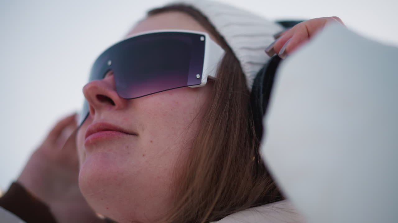 Close up of youthful spirit wearing over ear headphones under bright sun with beanie and jacket, hair glinting in light, gentle smile showing content connection to music and winter breeze excitement