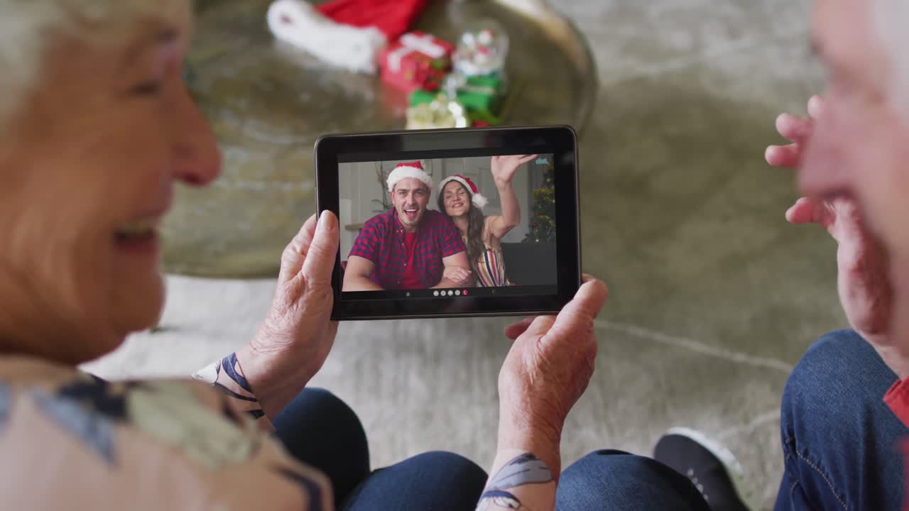 Anziana coppia caucasica che usa un tablet per una videochiamata natalizia con una coppia sorridente sullo schermo